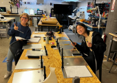 Two women give the camera a smile and thumbs-up from a work table. The table is covered in SteadyScrib tablets.